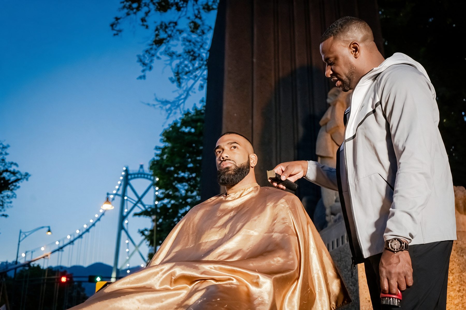 Junior cutting a client's hair at dusk beside the Lions Gate Bridge lions, the client draped in a gold cape